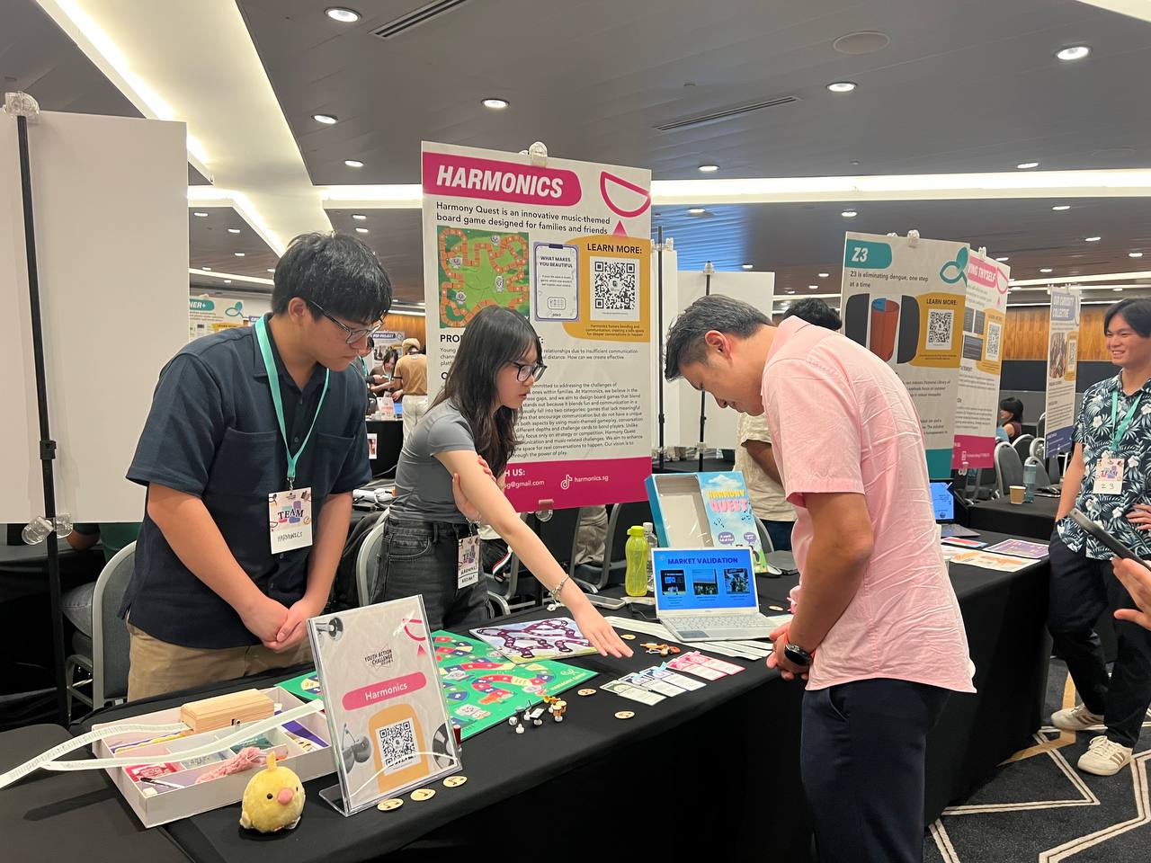 Shawn Huang at the Harmonics booth at Youth Plan 2025, looking at the Harmony Quest board with team members.